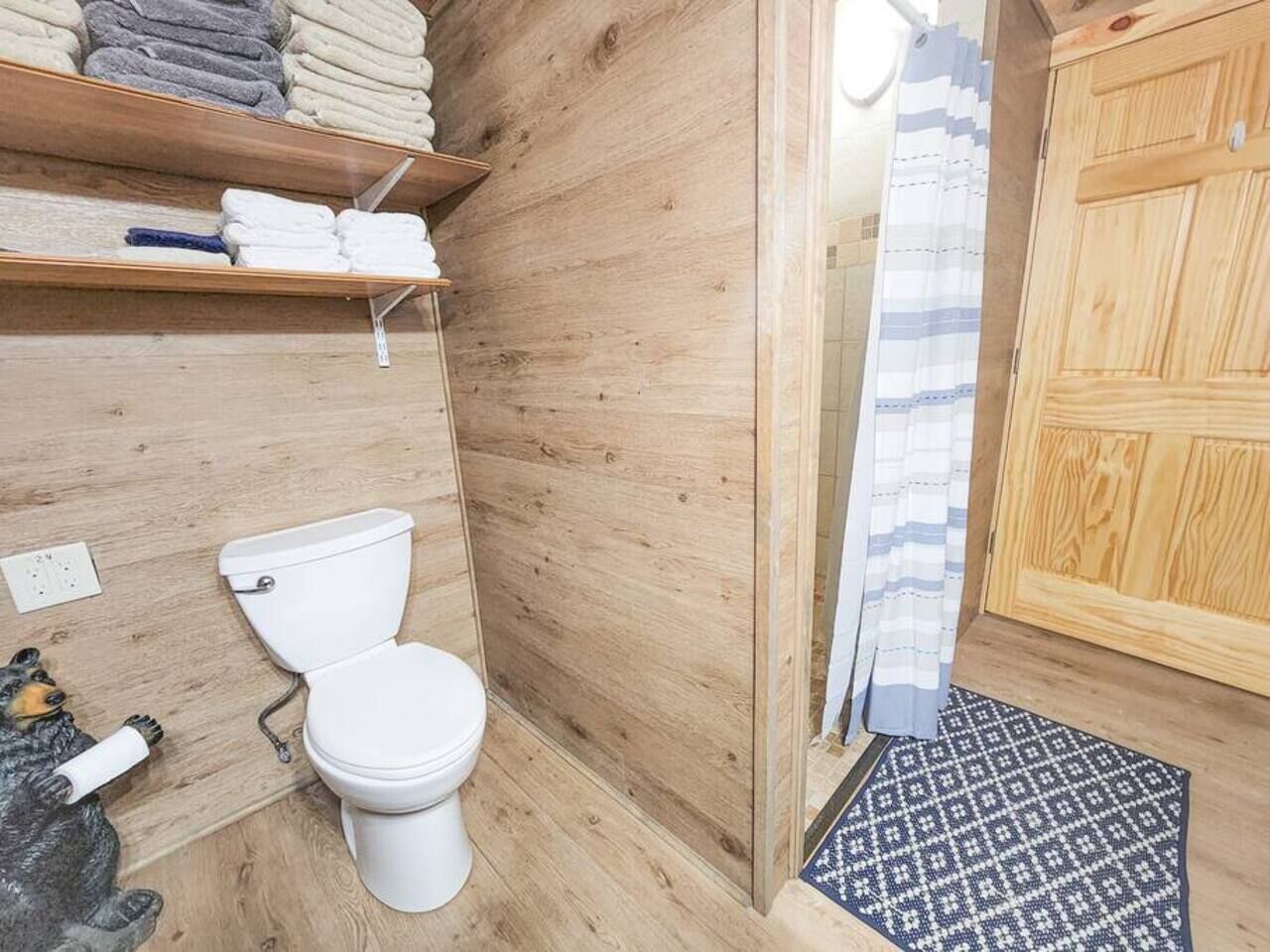 Un baño pequeño de madera con un inodoro, un estante con toallas dobladas y una alfombra estampada.