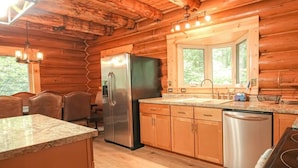 A log cabin kitchen with a stainless steel refrigerator, wooden cabinets, granite countertops, and a window with a view of greenery.