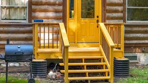 A log cabin with a yellow front door, a wooden staircase, and a barbecue grill.