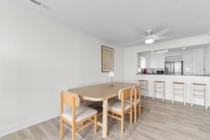 A dining area with a round table and four chairs, a ceiling fan, and a kitchen in the background.