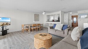 A modern living room with a dining area, kitchen, and a large window with a beach view.
