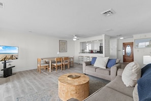 A modern living room with a dining area, kitchen, and a large window with a beach view.