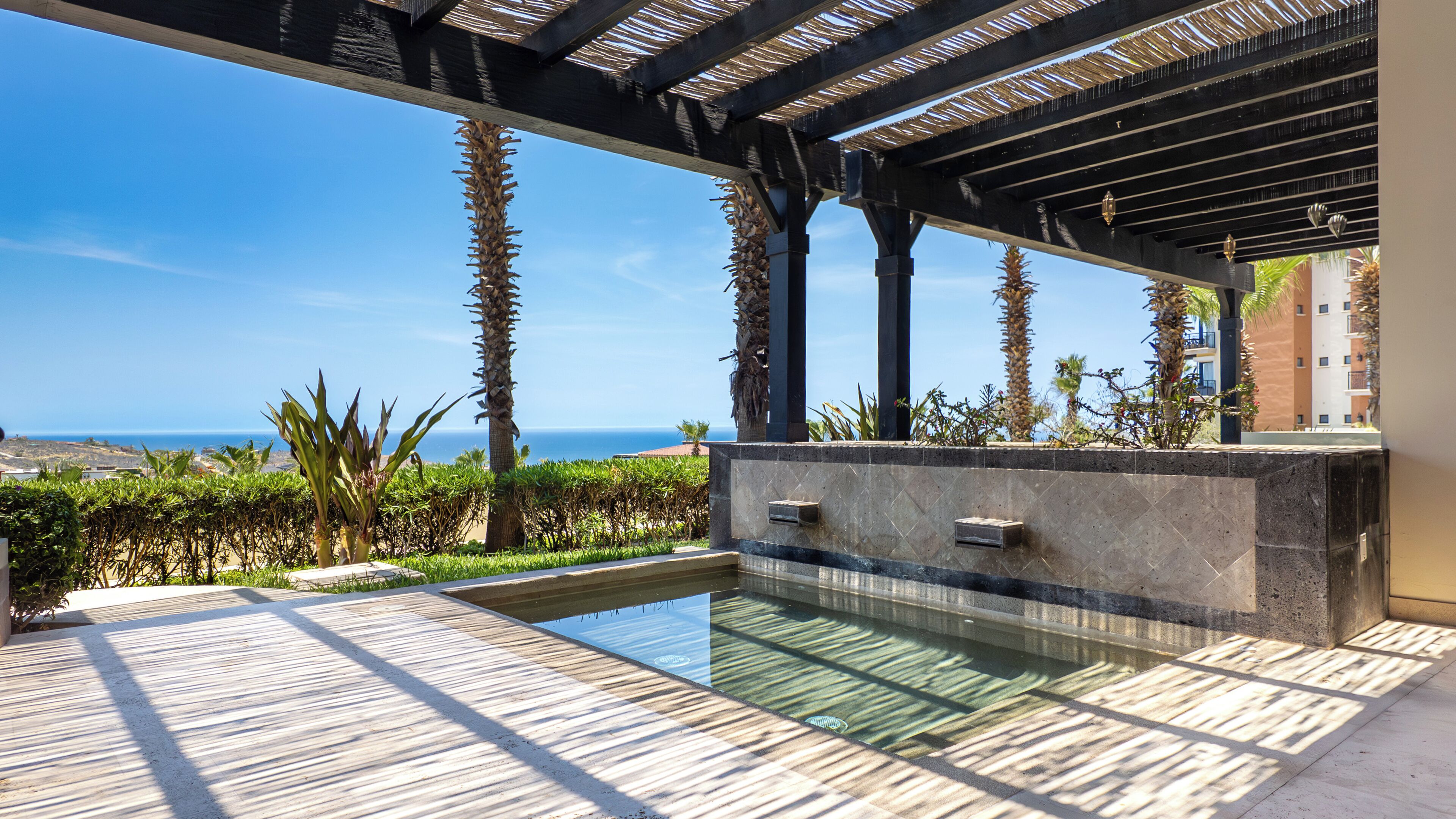 A pool area with a view of the ocean, surrounded by palm trees and a pergola structure.