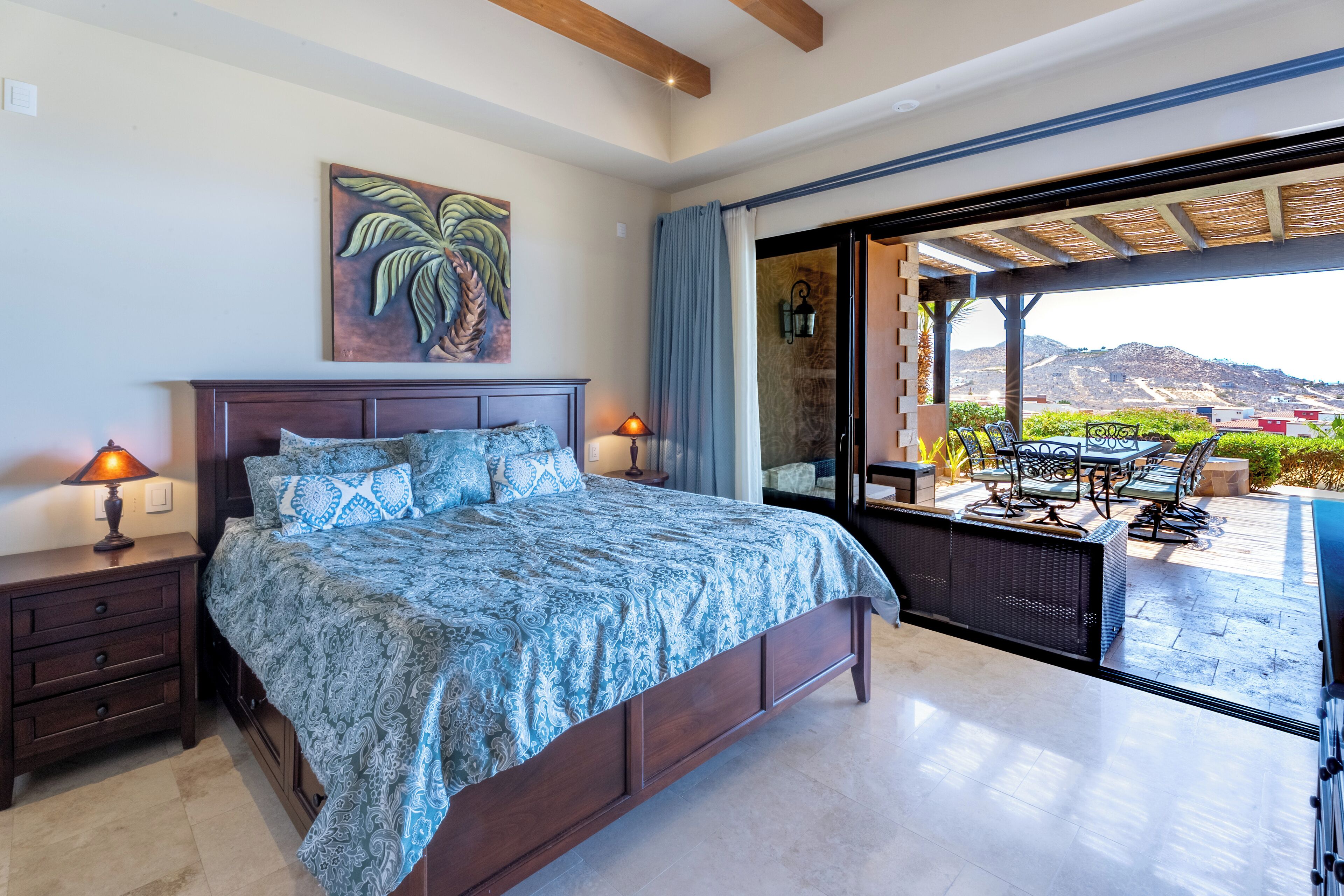 A bedroom with a large bed, a nightstand, a lamp, and a view of an outdoor patio with furniture.
