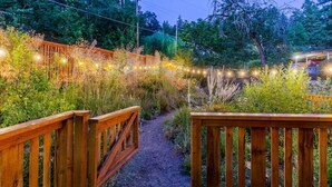A wooden pathway leading through a garden with string lights and dense foliage.