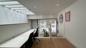 Ein moderner Büroraum mit einem langen weißen Tisch, schwarzen Bürostühlen und Holzboden. Eine Treppe führt zu einer anderen Ebene, und in der Nähe der Treppe steht ein Topfpflanze.
