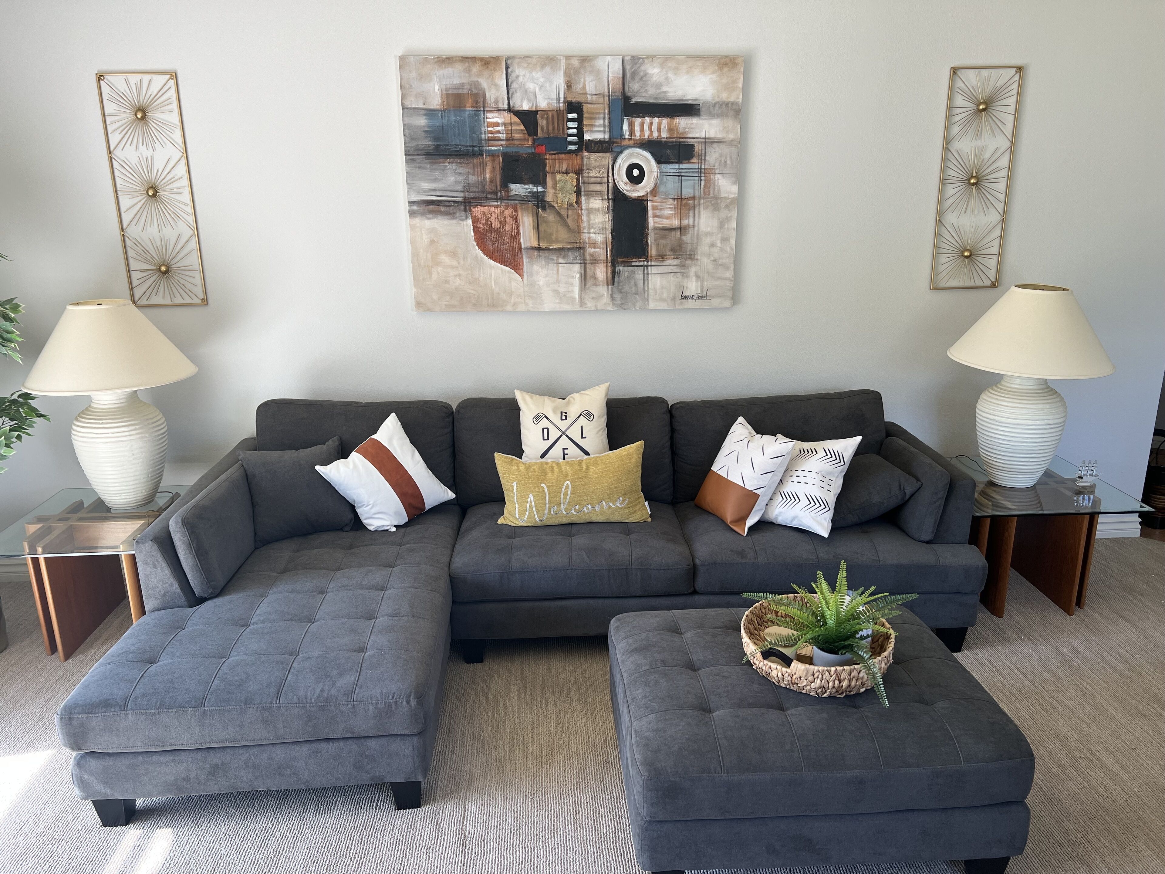 A modern living room with a grey sectional sofa, two side tables with lamps, a coffee table with a plant, and a large abstract painting on the wall.