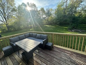 A deck with a wooden table and a cushioned seating area, surrounded by a wooden railing and a grassy yard.