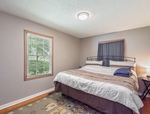 A bedroom with a single bed, a window with blinds, a ceiling light, and a small bedside table.
