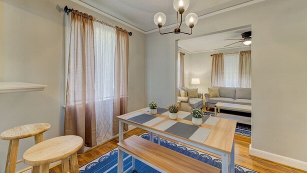 A modern living room with a wooden coffee table, two armchairs, and a sofa. There is a dining table with chairs, a chandelier, and a large mirror reflecting the room.