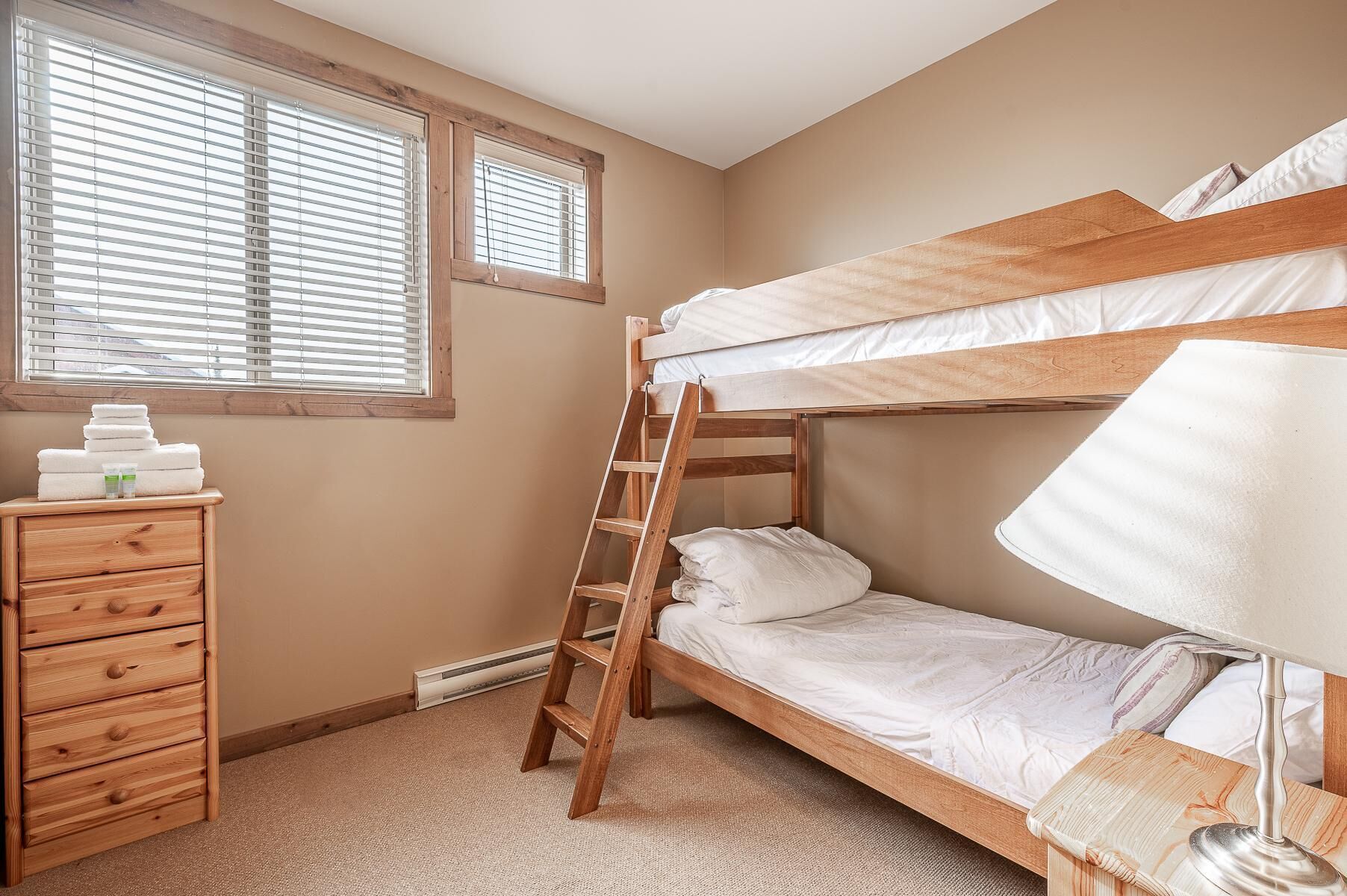 Dormitorio con literas, una escalera de madera, mesita de noche con lámpara y ventana con persianas.
