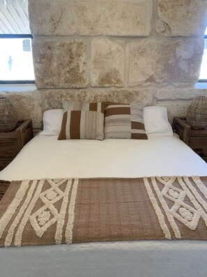 A bed with a white mattress, brown and white striped pillows, and a brown and white patterned throw. The bed is against a stone wall. - The Rock Stack: Ruggedly luxurious 3 bedroom, 3 bathroom on the Llano River (Llano)