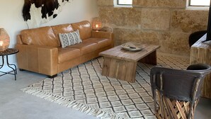 A cozy living room with a stone wall, a leather sofa, a wooden coffee table, a woven rug, and a unique chair. - The Rock Stack: Ruggedly luxurious 3 bedroom, 3 bathroom on the Llano River (Llano)