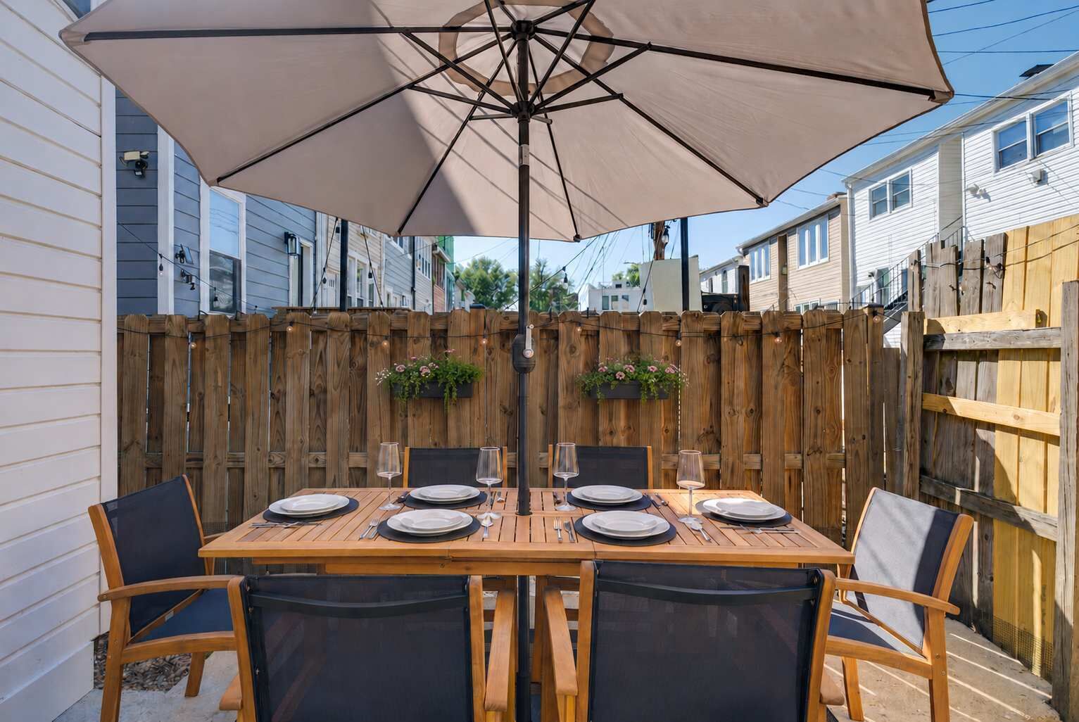 A wooden table set for six with a large umbrella, surrounded by chairs, in a backyard with a wooden fence and houses in the background.