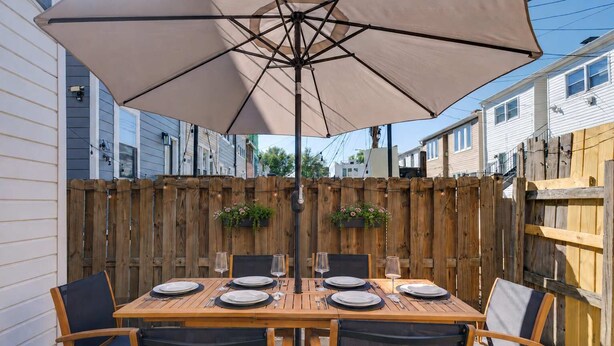 Une table en bois, dressée pour six personnes, est placée sous un grand parasol, entourée de chaises, dans un jardin avec une clôture en bois et des maisons en arrière-plan.