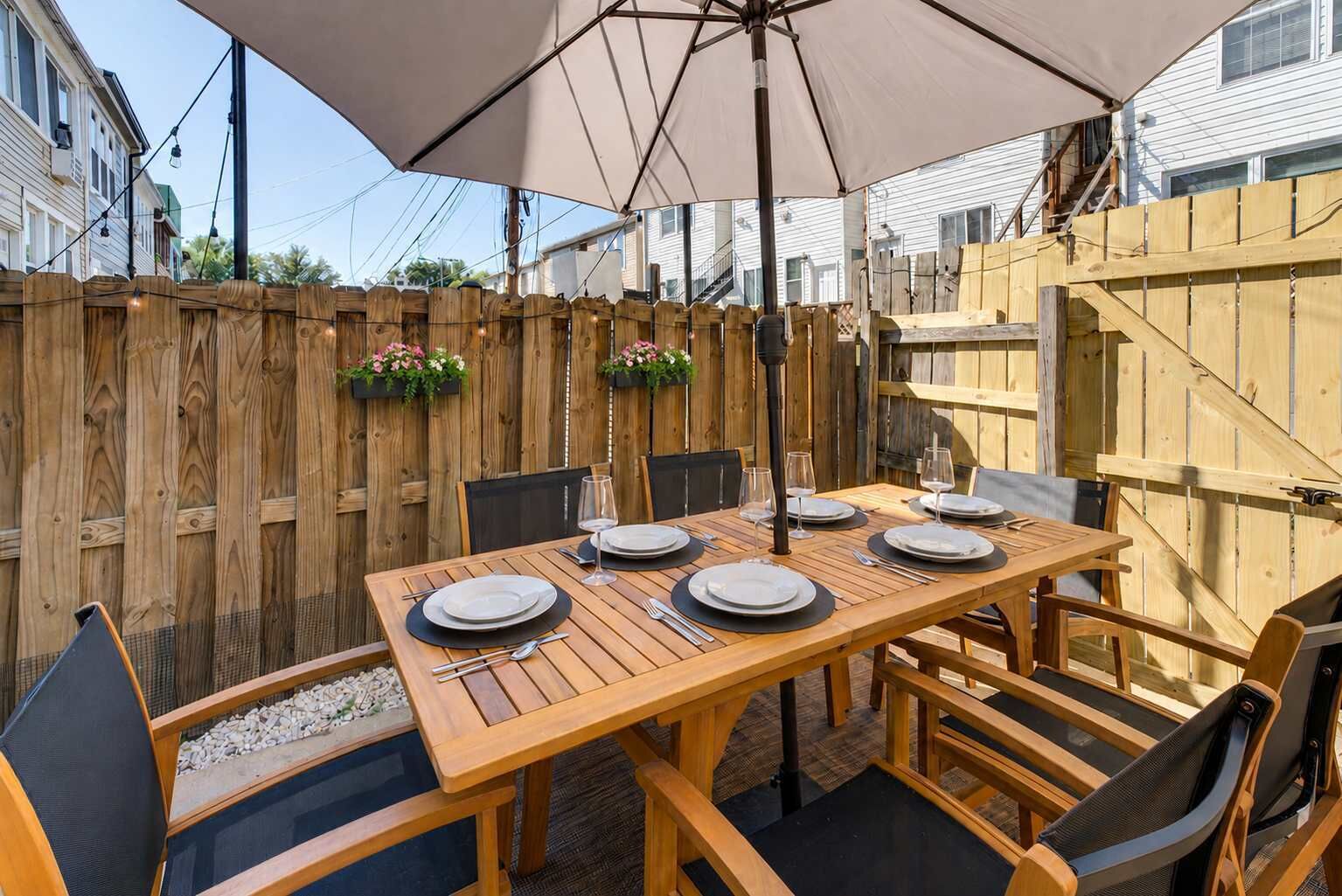 An outdoor wooden table set with plates and glasses, surrounded by chairs, under a large umbrella.