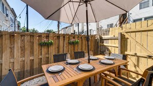 Une table en bois installée à l’extérieur, avec des assiettes et des verres, entourée de chaises, et protégée par un grand parasol.