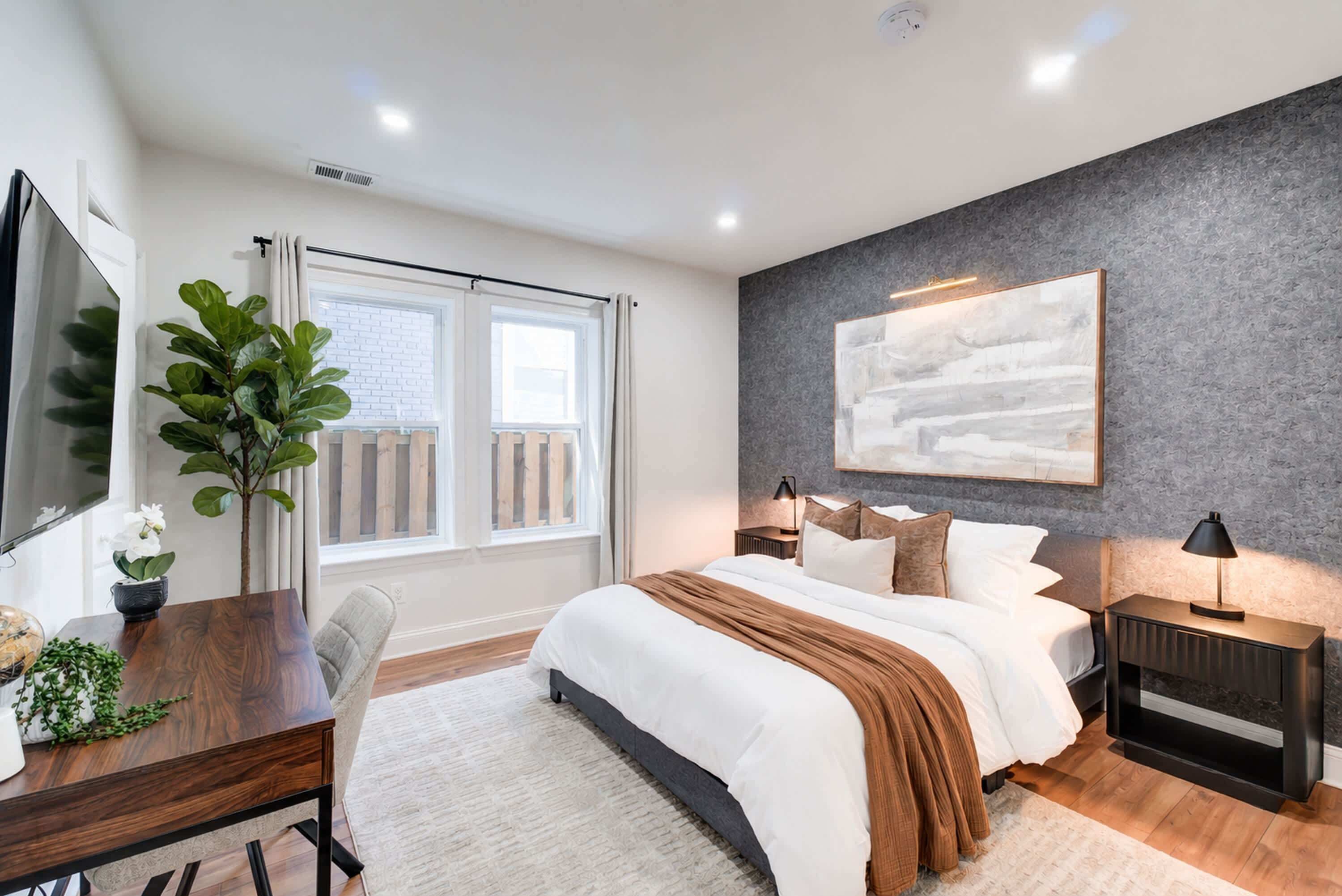 A modern bedroom with a large bed, a desk, a chair, a potted plant, and a window with curtains.