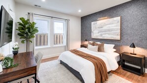 Une chambre à coucher moderne avec un grand lit, un bureau, une chaise, une plante en pot et une fenêtre avec des rideaux.