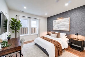Un dormitorio moderno con una cama grande, un escritorio, una silla, una planta en maceta y un ventanal con cortinas.