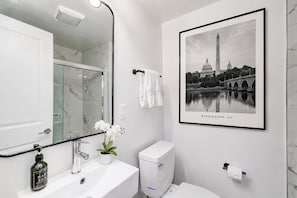 Un baño con lavamanos de mármol, un inodoro, un cuadro enmarcado del Monumento a Washington y una orquídea en maceta.