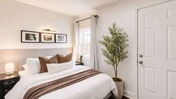 A neatly arranged hotel room with a bed, bedside table, lamp, framed pictures, a potted plant, and a white door.