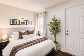 A neatly arranged hotel room with a bed, bedside table, lamp, framed pictures, a potted plant, and a white door.