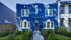 Un bâtiment à deux étages, à l’extérieur bleu, avec une clôture en bois et un jardin bien entretenu agrémenté de haies.