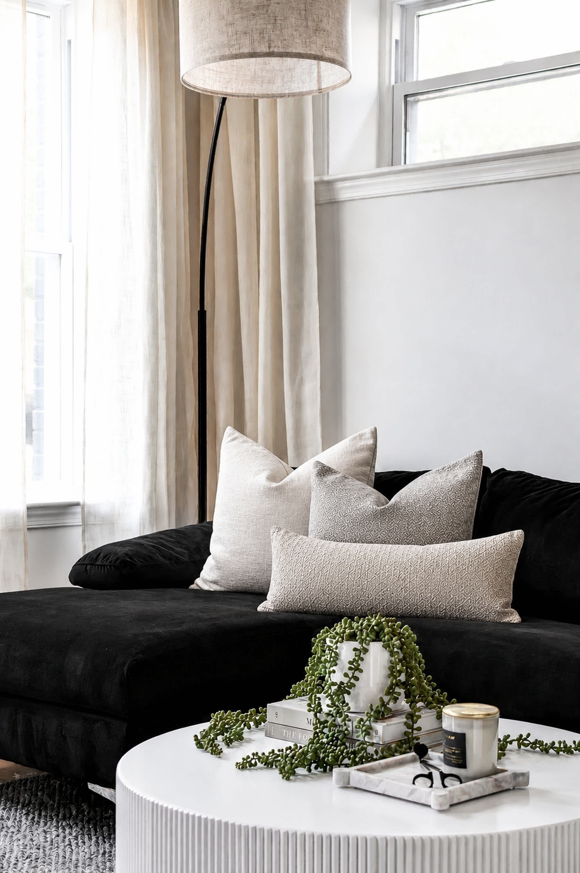 A modern living room with a black sofa, white cushions, a white coffee table with decorative items, and a tall floor lamp.