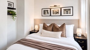 Een net opgemaakt bed met een hoofdbord, twee nachtkastjes met lampen, en drie ingelijste foto’s aan de muur.