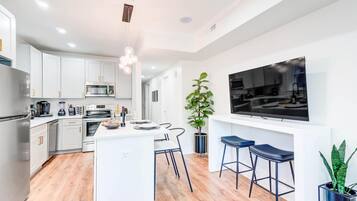 A modern kitchen with white cabinets, a central island, bar stools, a large TV, and a potted plant.