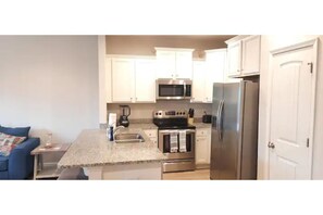 Compact kitchen with stainless steel appliances, granite countertop, and a small island with a sink, adjacent to a cozy living area with a sofa and coffee table.