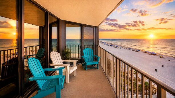 A balcony with two chairs, a table, and a view of the beach at sunset. - Gulf-View Balcony at Edgewater Resort (Panama City Beach)