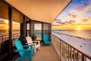 A balcony with two chairs, a table, and a view of the beach at sunset.