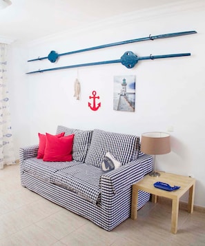 Ein kariertes Sofa mit roten Kissen, ein kleiner Tisch mit einer Lampe und maritime Dekoration, darunter ein Anker und ein Leuchtturm-Foto.