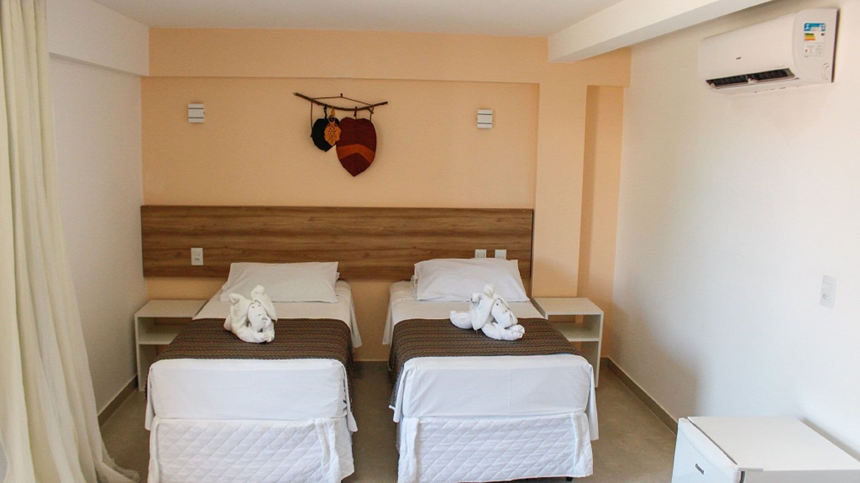 A hotel room with two beds, each with a white quilted mattress and pillows. There is a wooden headboard, a wall-mounted air conditioning unit, and a small white cabinet.