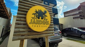 Opastekyltti, jossa lukee ”Jardins Tropical”, ja jossa on trooppisen talon kuva. Taustalla näkyy moderni rakennus ja edessä pysäköity auto.