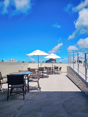 Un patio al aire libre con sillas y mesas de mimbre, sombrillas blancas y vistas a edificios al fondo.