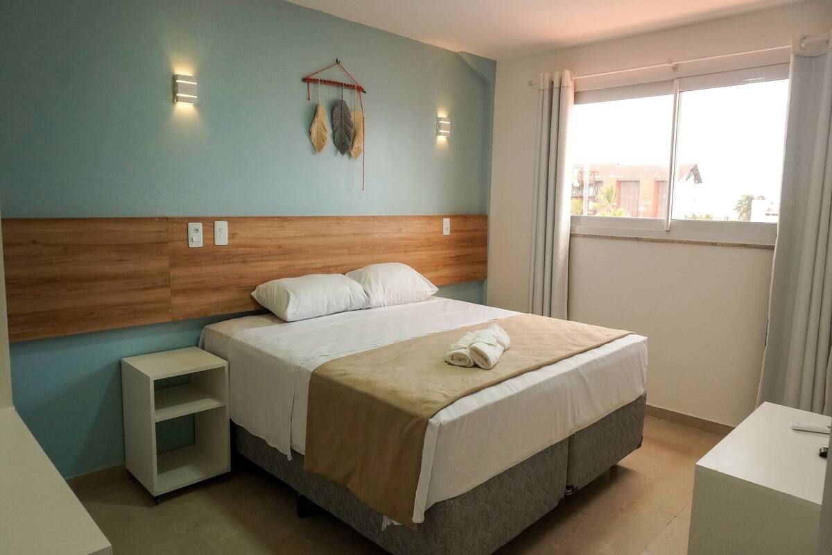 A neatly made bed with a beige blanket and white pillows, a bedside table, a small shelf, and a wall-mounted light fixture.