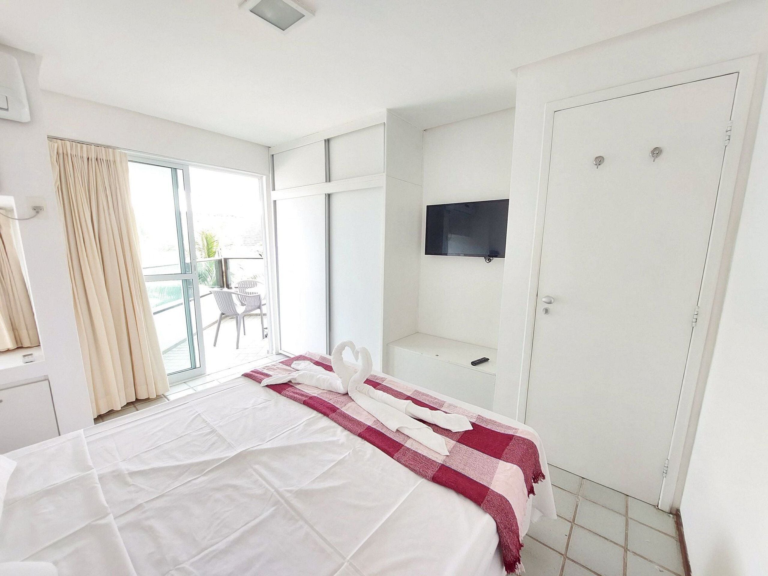 A hotel room with a bed, a TV, a balcony with chairs, and a door.