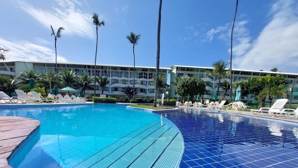 A swimming pool with tiled flooring, surrounded by lounge chairs and palm trees, with a building in the background. - Ancorar Flat Resort (Ipojuca)