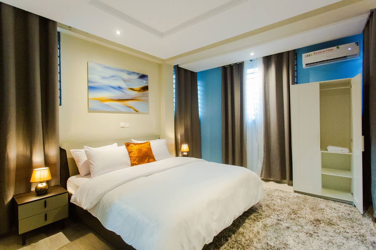 Une chambre d’hôtel avec un grand lit, une table de chevet avec une lampe, une armoire et un climatiseur fixé au mur.