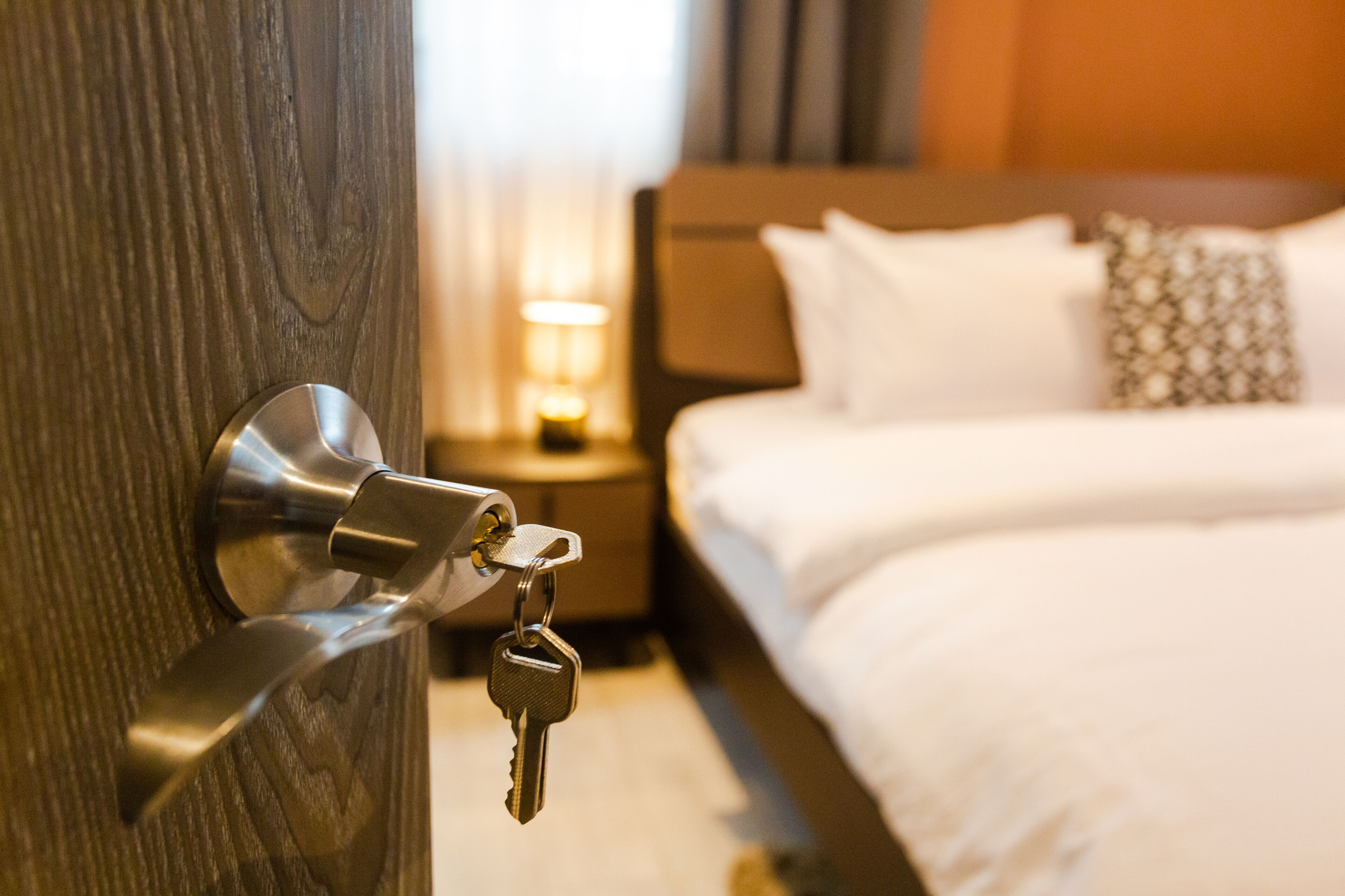 A hotel room with a wooden door, a bed with white bedding, and a nightstand with a lamp.
