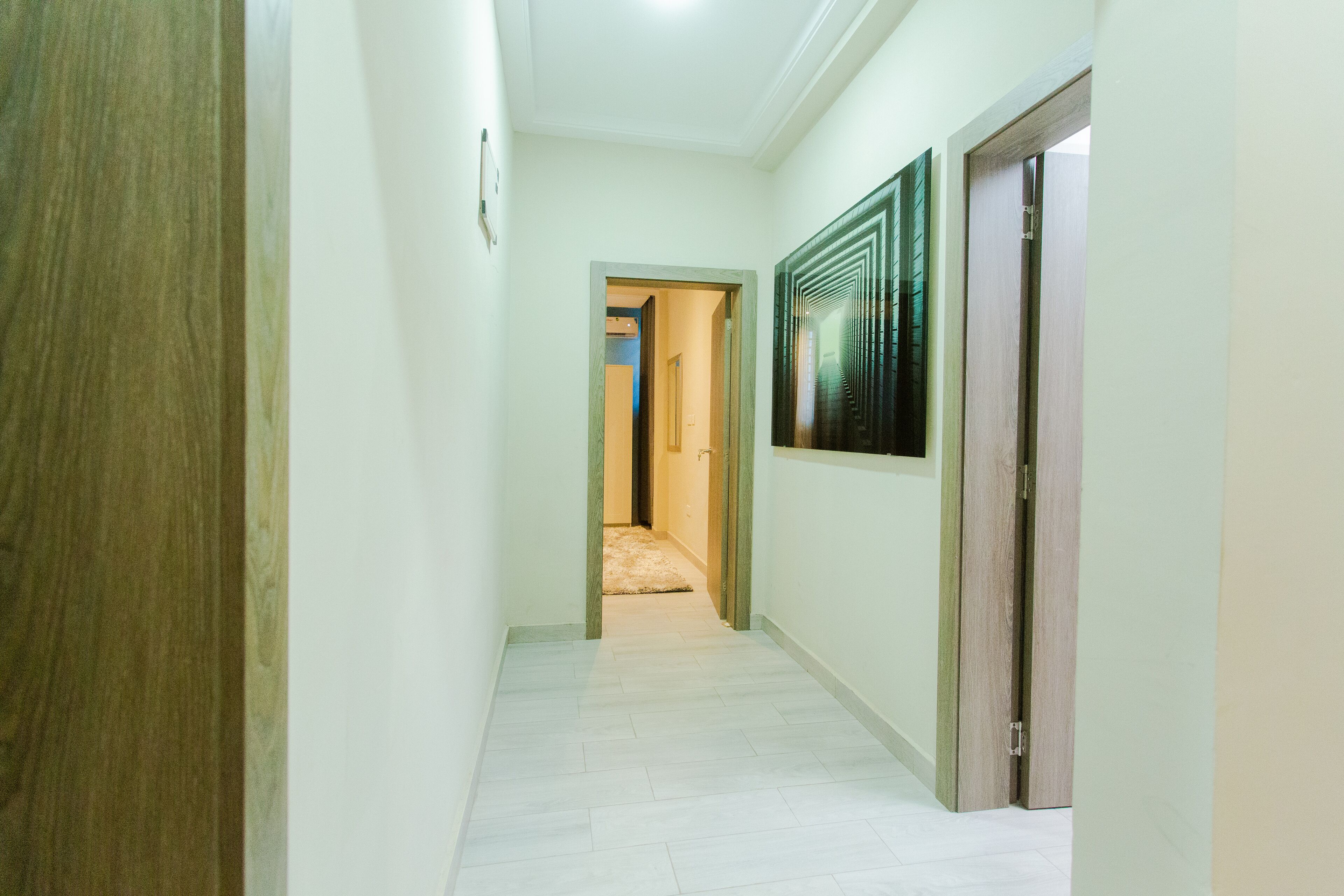 Un couloir avec une porte en bois sur la gauche, une œuvre d’art encadrée sur la droite et une zone recouverte de moquette au fond.