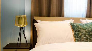 A neatly made bed with white and green pillows, a small bedside table with a lamp, and a nightstand with drawers.