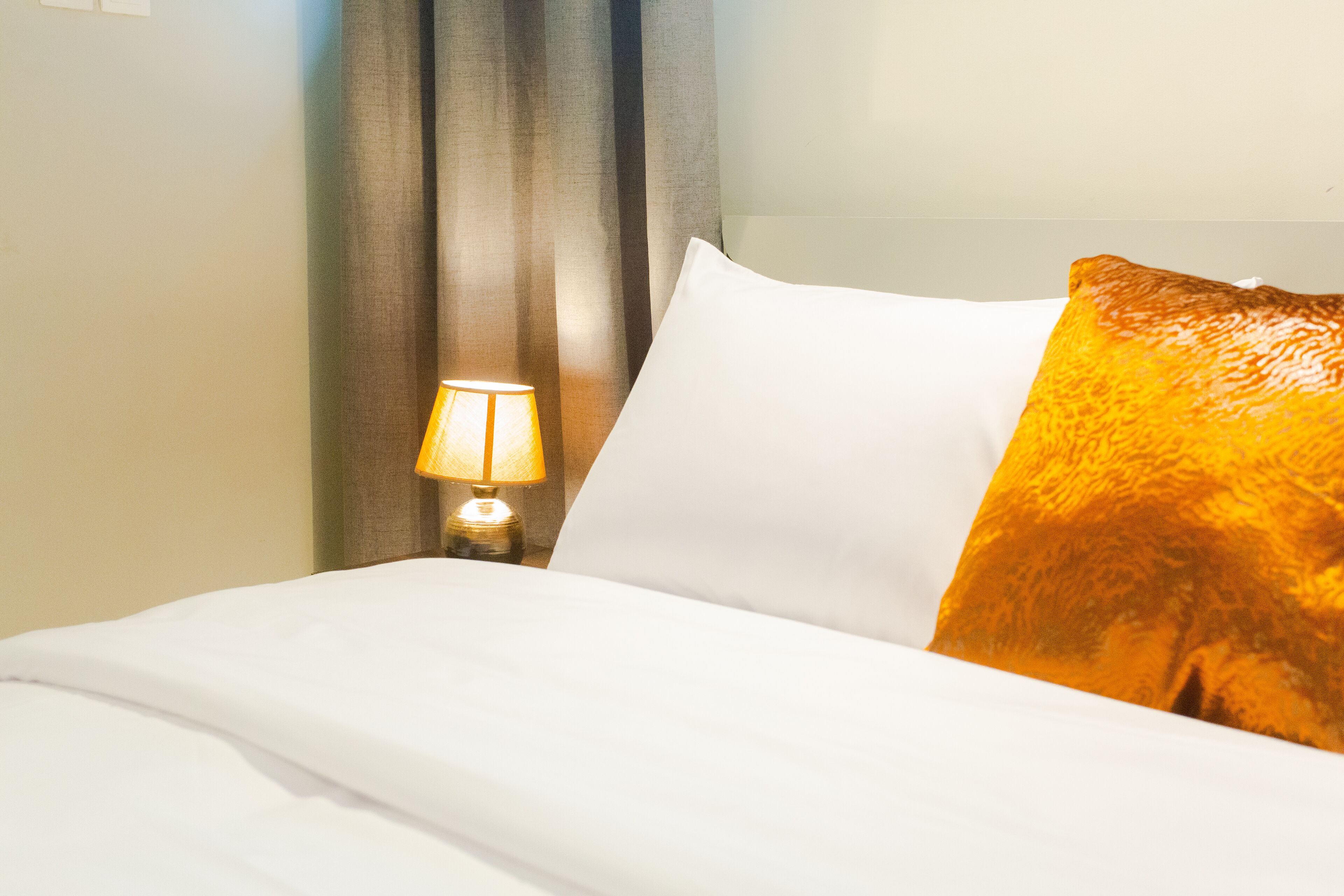 A neatly made bed with white linens, a single white pillow, and a decorative orange pillow. A small table lamp with a white shade is visible on the nightstand beside the bed.