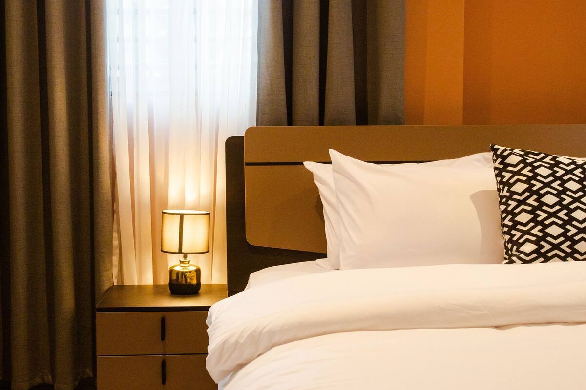 Une chambre d’hôtel avec un lit, une table de chevet avec une lampe, une fenêtre avec des rideaux et un plancher en bois.