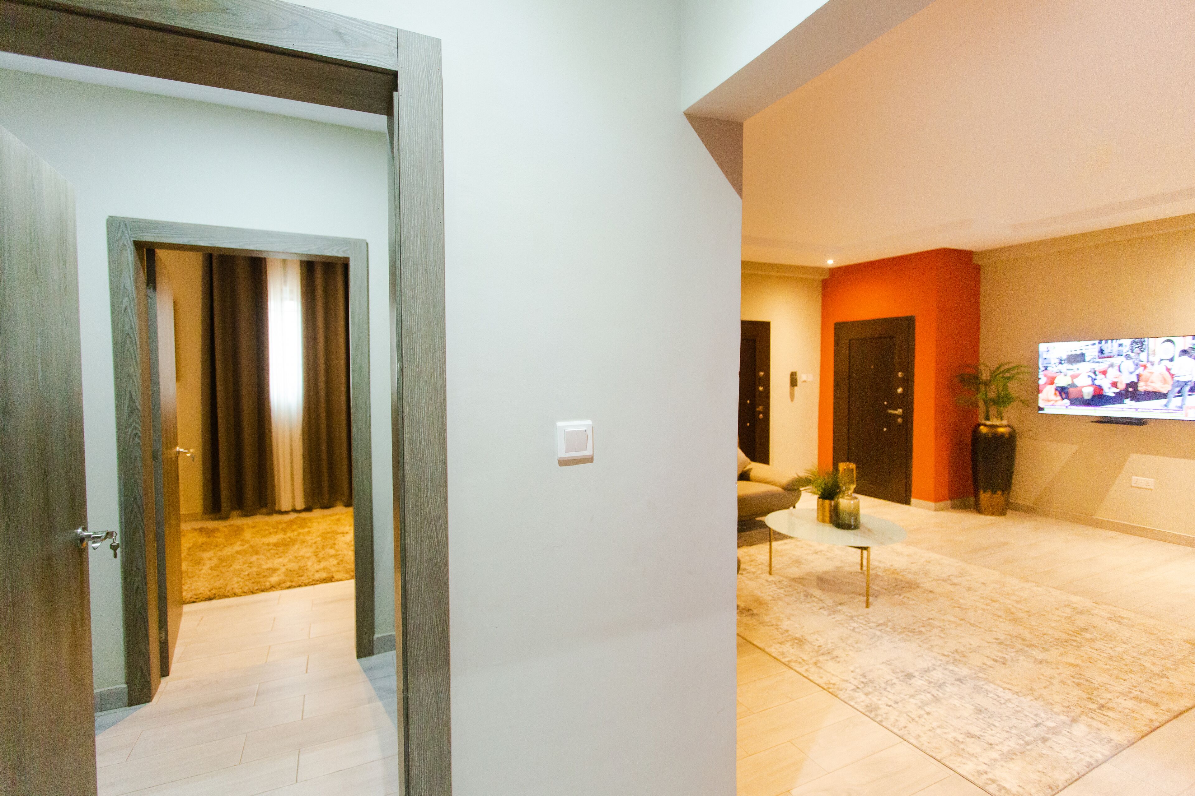 A modern interior with a wooden door, a hallway leading to a living area, and a flat-screen TV mounted on the wall.