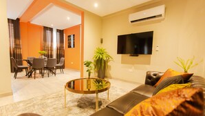 Una sala de estar moderna con un sofá, una mesa de centro y un televisor montado en la pared. Se observa un comedor con sillas y una mesa al fondo.