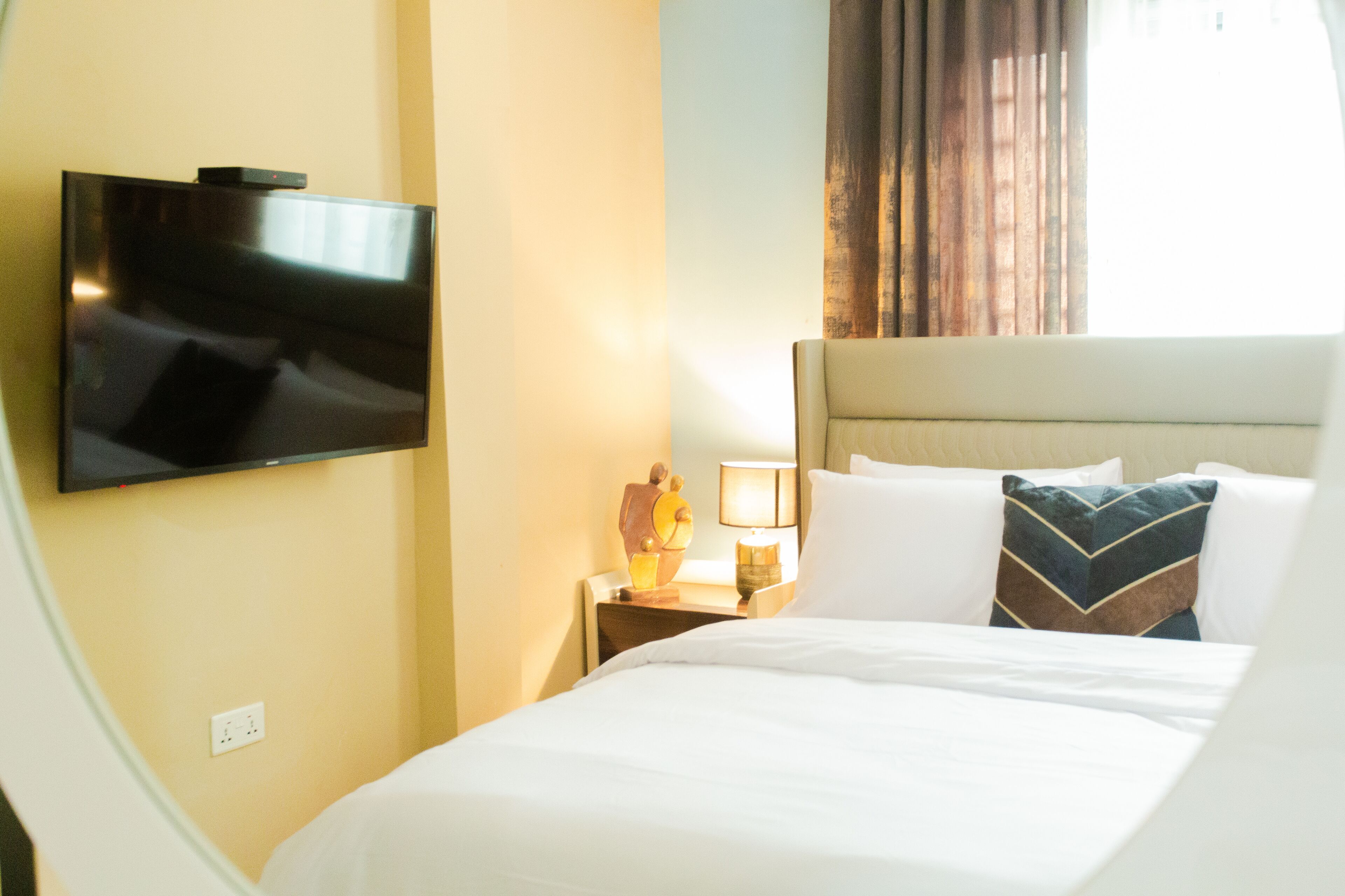 Una habitación de hotel con un televisor de pantalla plana montado en la pared, una mesita de noche con lámpara y una cama con ropa blanca y almohadas decorativas.